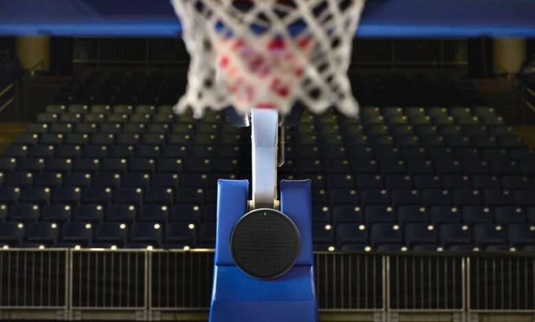 Close-up of a basketball hoop with a speaker mounted on the backboard, overlooking rows of empty seats in an arena.