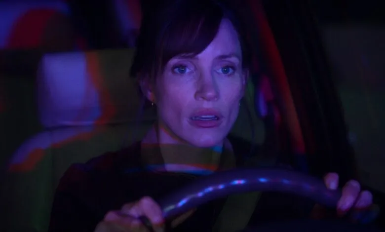 Close-up of a woman with a worried expression driving at night, bathed in purple and blue light.