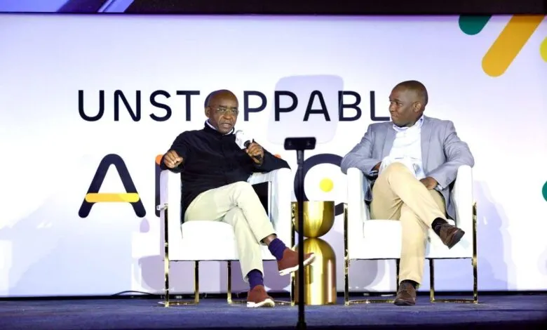 Two men in business attire sit on stage at an AI conference, one speaking into a microphone.