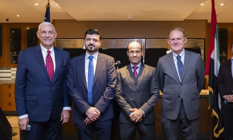 Four men in suits stand together, with UAE and US flags in the background.