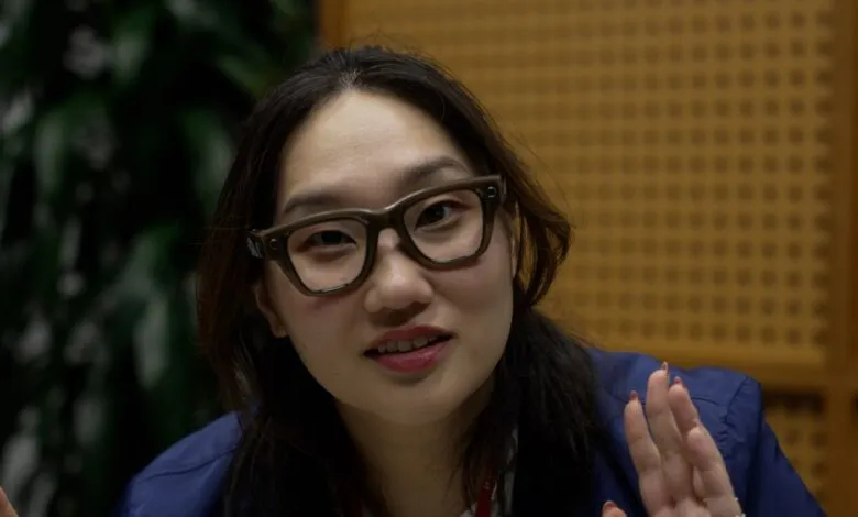 Close-up of a young woman with dark hair and glasses, speaking, possibly in an interview setting.