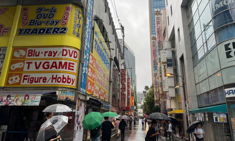 Rainy day in Akihabara, Tokyo, people with umbrellas walking past shops selling games, anime figures, and electronics.