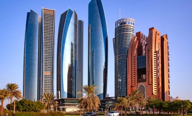 Modern skyscrapers in Abu Dhabi, UAE, reflecting sunlight. Palm trees and a car are in the foreground.