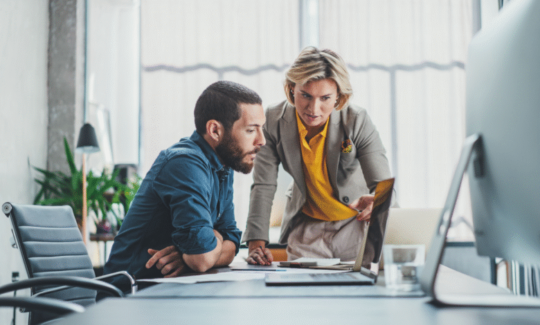 Two colleagues collaborate at a modern office desk, reviewing documents on a laptop.