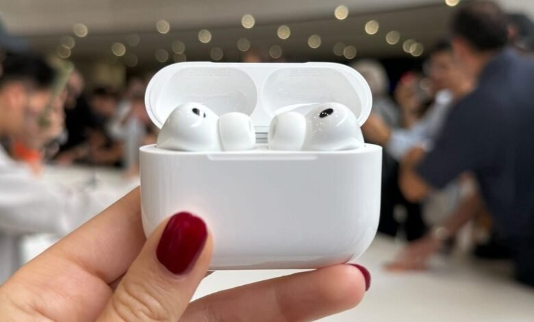 White AirPods Pro in their charging case, held in a hand with dark red nails. Blurred background shows people.