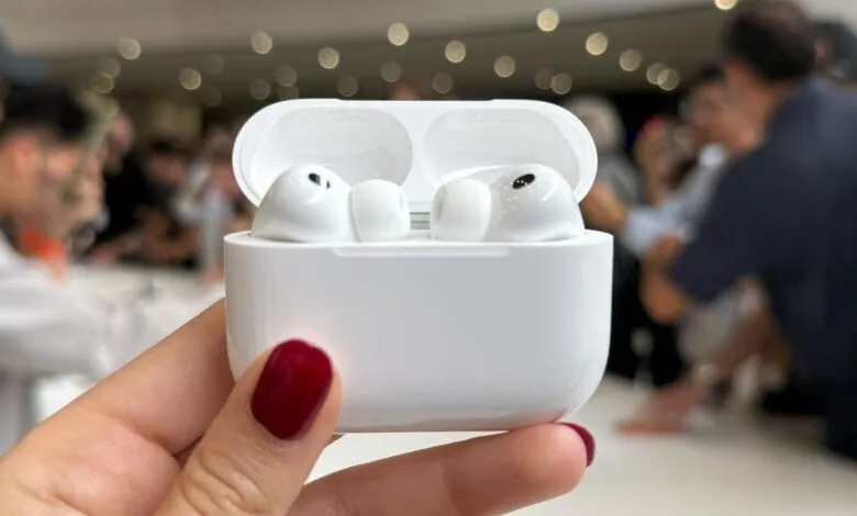 White AirPods Pro in their charging case, held in a hand with dark red nails. Blurred background shows people.