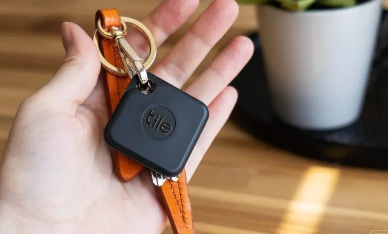 Hand holding a black Tile tracker attached to keys on an orange leather strap.