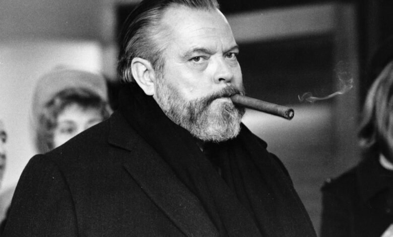 Orson Welles, a distinguished man with a beard, smokes a cigar in a black and white portrait.