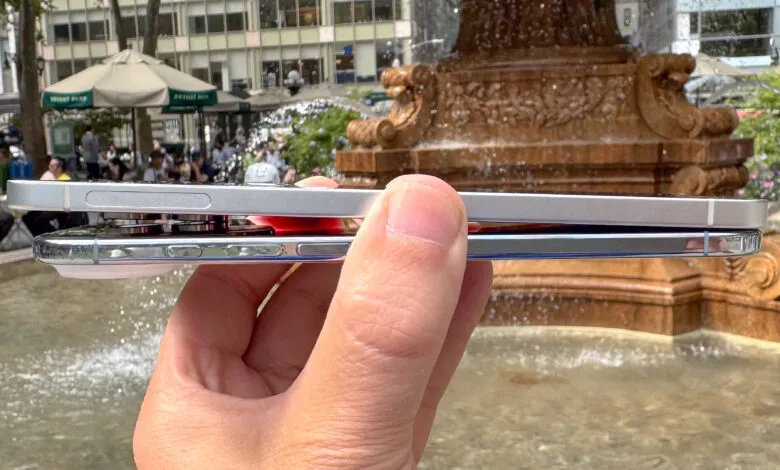 Close-up of a hand holding two smartphones stacked, with a fountain and city buildings in the background.
