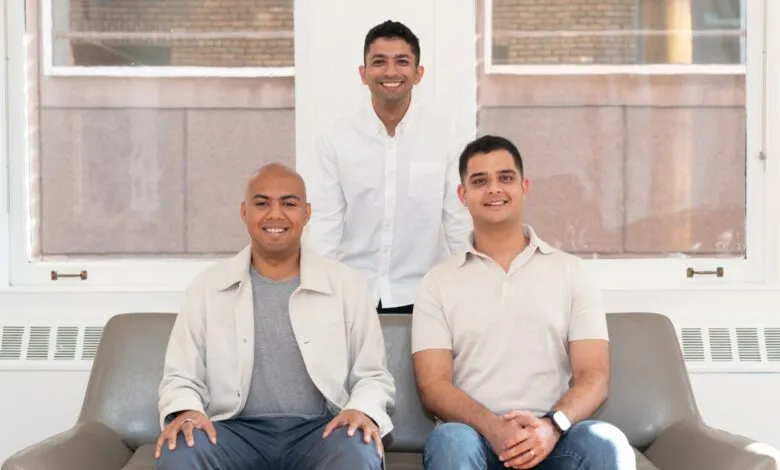 Three smiling men, two seated and one standing, pose for a professional portrait against a bright window.