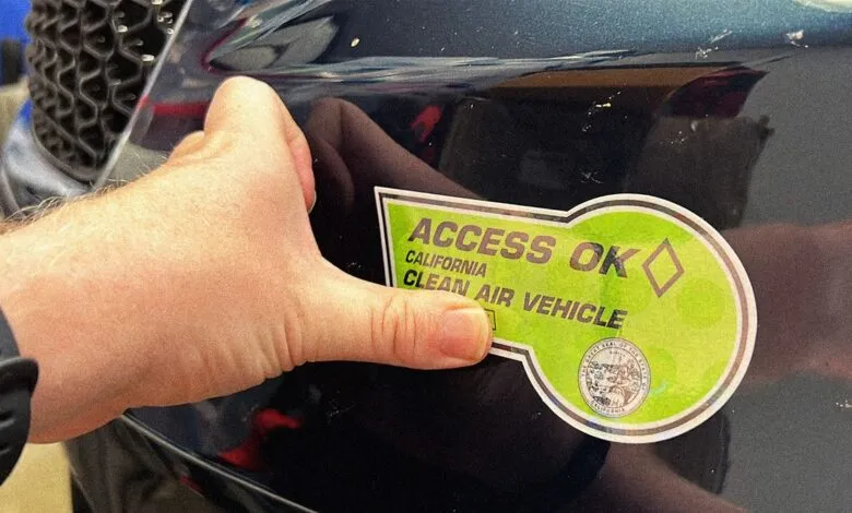 Close-up of a hand holding a California Clean Air Vehicle sticker on a dark-colored car.