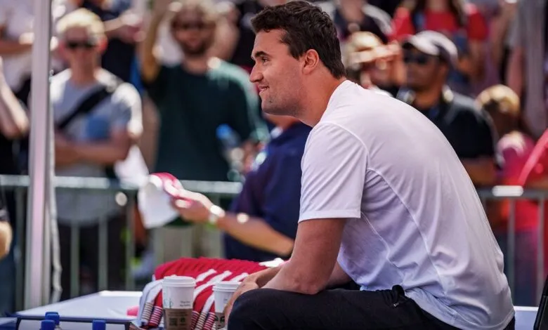 Charlie Kirk, conservative commentator, sits at a table outdoors, surrounded by a crowd.