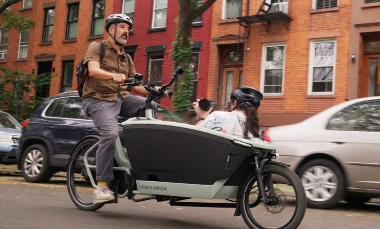 Man rides an Urban Arrow cargo ebike with a photographer in the cargo box, on a city street.