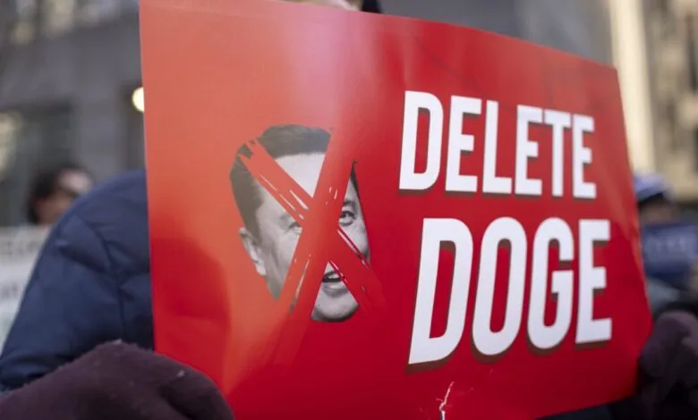 Protester holds a red sign with Elon Musk's face crossed out and the words 'DELETE DOGE'.