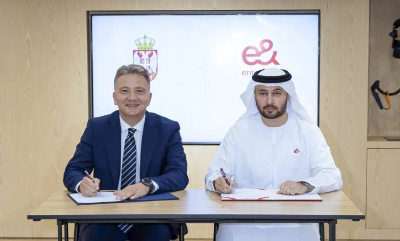 Two men in formal attire signing a document at a table. A screen displays the Serbian coat of arms and a logo.