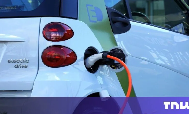 White electric car charging with an orange cable. The car's rear is visible, showing its taillights and 'electric drive' emblem.