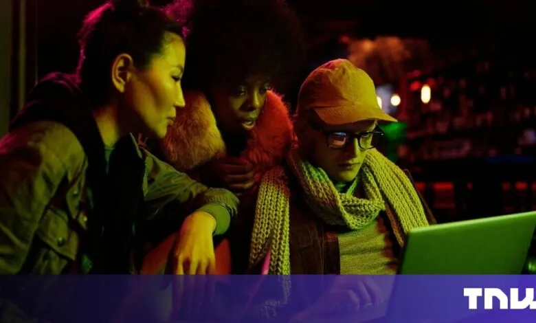 Three young adults huddle around a laptop at night, illuminated by vibrant neon lights.
