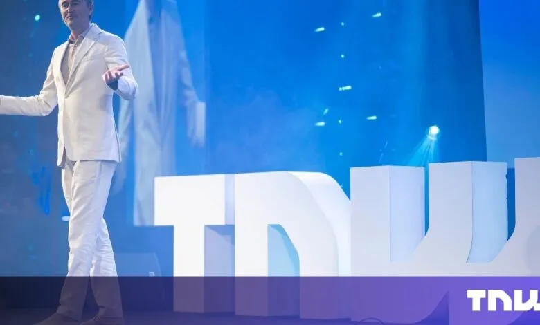 Man in a white suit speaking on stage with large white letters spelling TNW behind him.