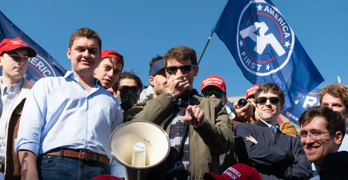A group of young men at a political rally, some wearing "Make America Great Again" hats, holding a megaphone and flags.