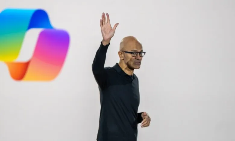 Satya Nadella, Microsoft CEO, waves at a presentation with a colorful logo in the background.
