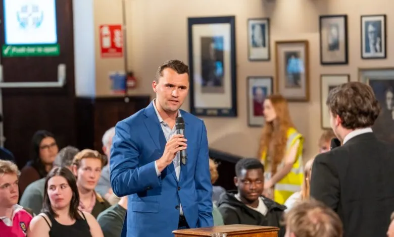 Man in blue suit speaks at a microphone to a diverse audience in a room with framed portraits.