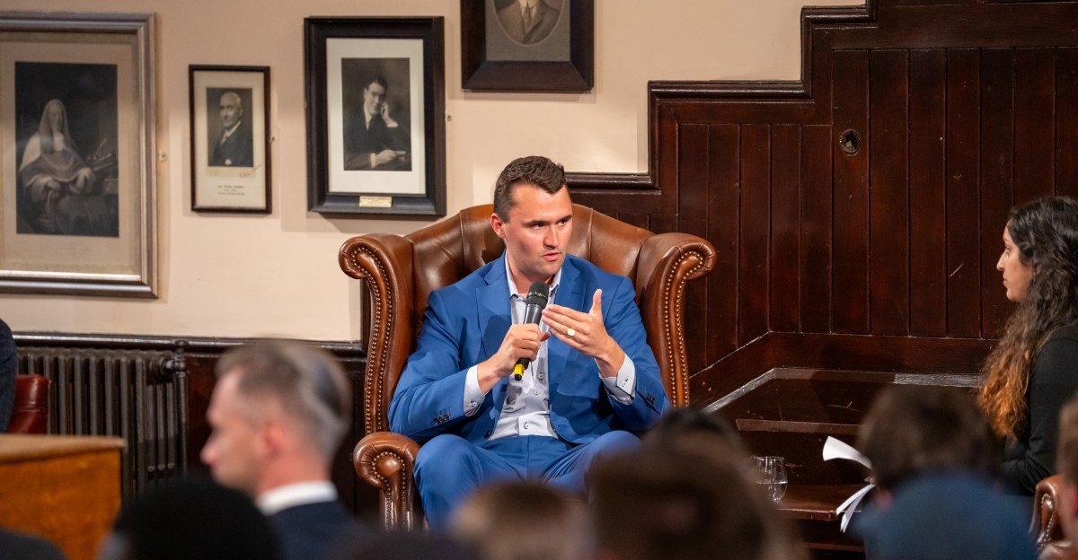 Man in blue suit speaks at a panel discussion, holding a microphone. He's seated in a leather armchair in a wood-paneled room.