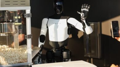 Tesla Optimus robot waves near a popcorn machine while a person films it with a smartphone.