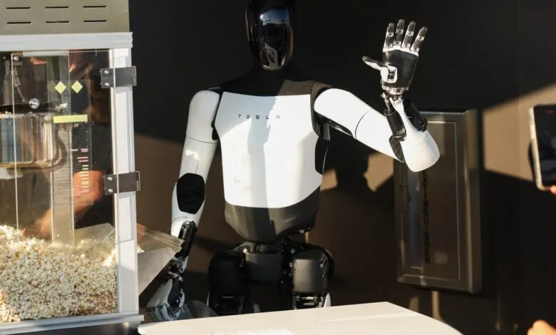 Tesla Optimus robot waves near a popcorn machine while a person films it with a smartphone.