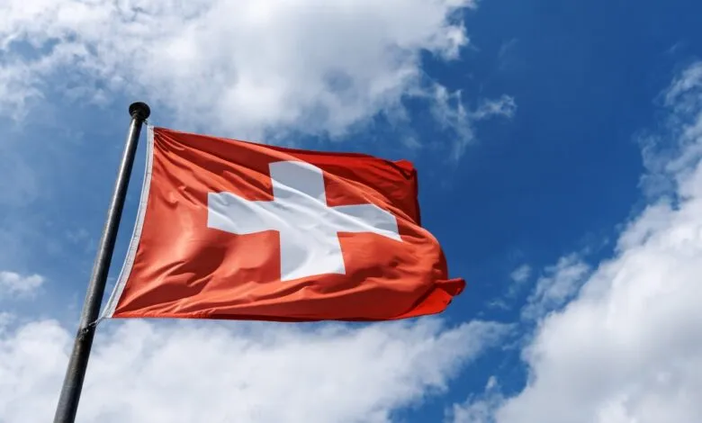 Swiss flag waving against a bright blue sky with fluffy white clouds.