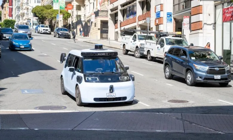 A Waymo self-driving car navigates a San Francisco street, surrounded by other vehicles and pedestrians.