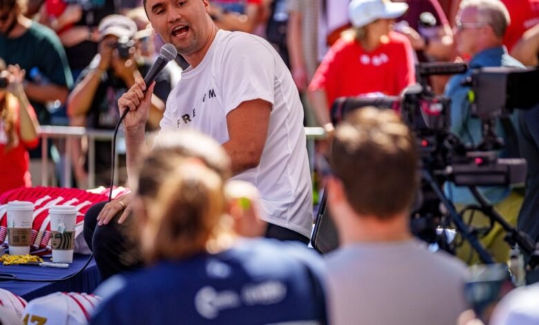 Tim Tebow speaks at an outdoor event, holding a microphone. A crowd surrounds him, some holding cameras.