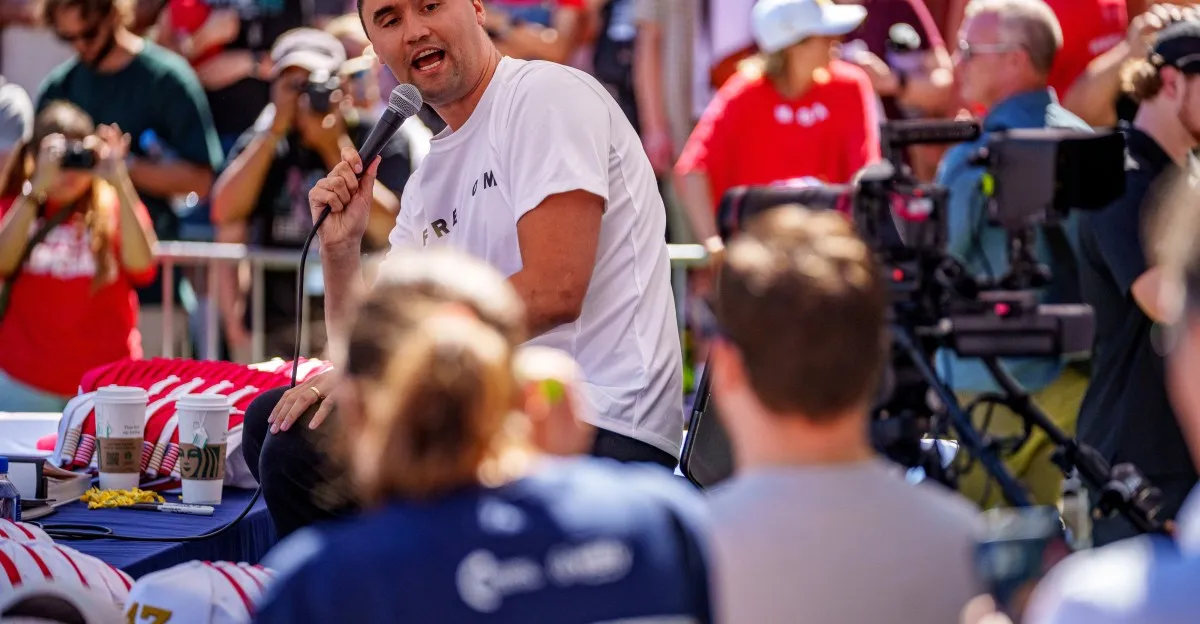 Tim Tebow speaks at an outdoor event, holding a microphone. A crowd surrounds him, some holding cameras.