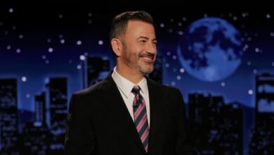 Jimmy Kimmel, smiling, in a suit and tie, on a late-night talk show set with a city skyline backdrop.