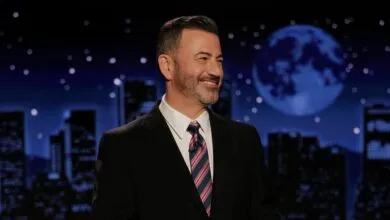 Jimmy Kimmel, smiling, in a suit and tie, on a late-night talk show set with a city skyline backdrop.