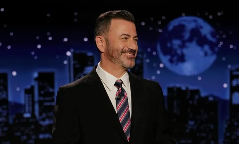 Jimmy Kimmel, smiling, in a suit and tie, on a late-night talk show set with a city skyline backdrop.