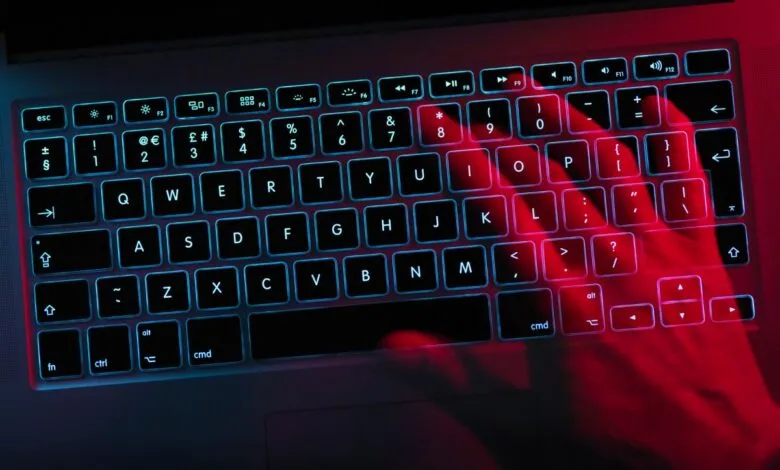 Close-up of a hand typing on a backlit laptop keyboard glowing red and blue. Cybersecurity or late-night work concept.