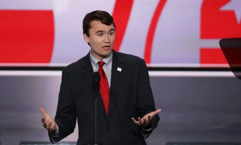 Young man in suit and red tie speaks at a podium, gesturing with hands. Red and white background.