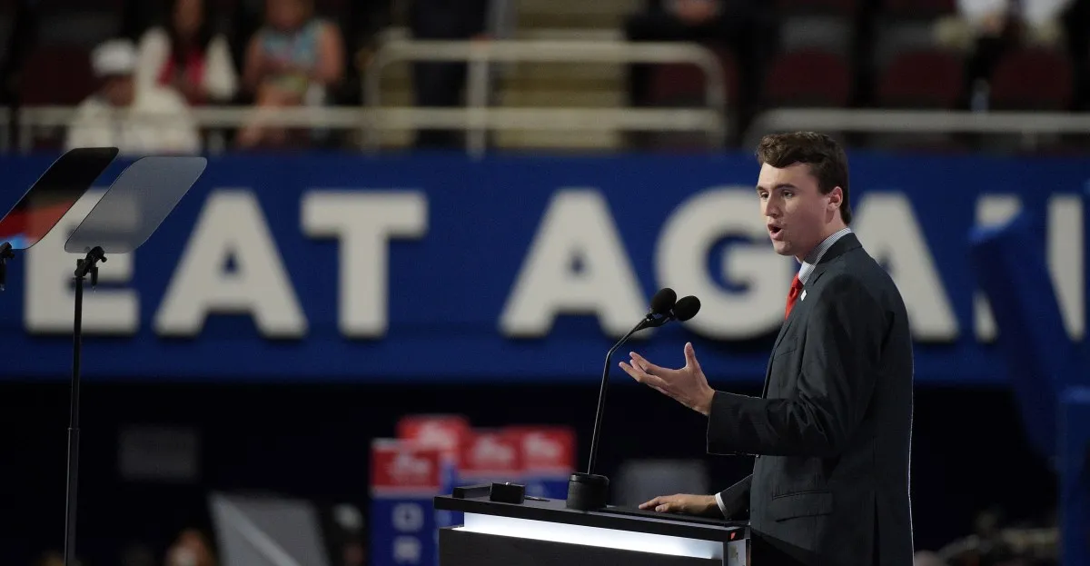 Young man in suit speaks at a podium, a large 'EAT AGAIN' sign blurred in the background.