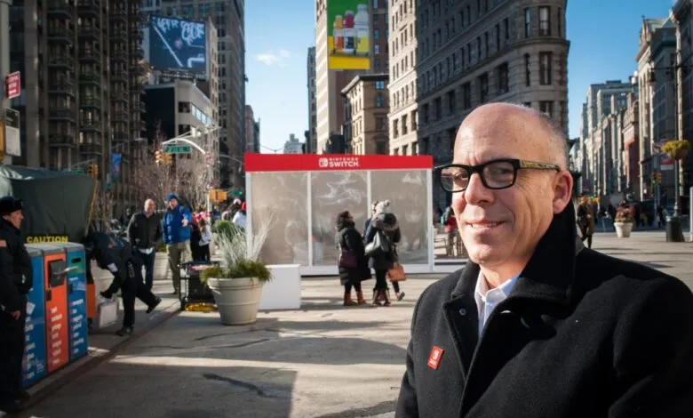 Man in glasses and black coat smiles at camera, with Nintendo Switch display and city street in background.