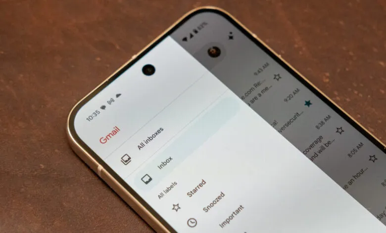 Gmail app on a smartphone showing inbox, starred emails, and other options. The phone rests on a brown surface.
