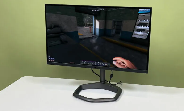 A dark gray computer monitor displays a first-person video game scene. A hand interacts with the game's interface.