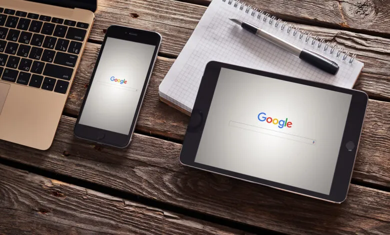 Google search displayed on a phone and tablet, alongside a laptop and notepad on a wooden desk.