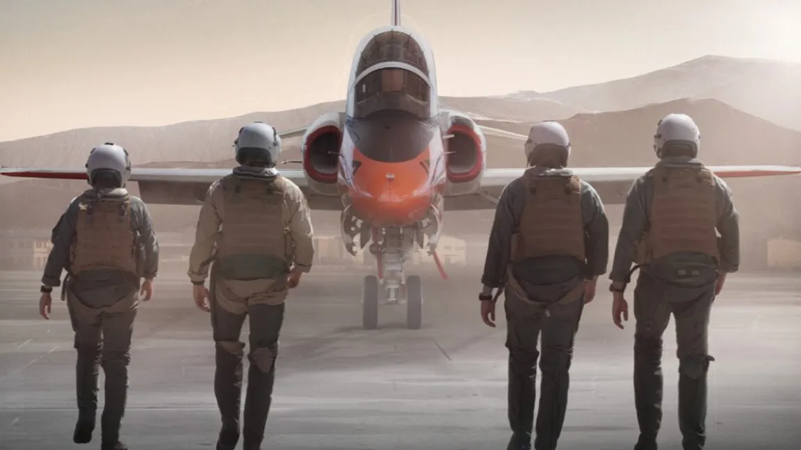 Four pilots in flight suits walk towards a military jet on a desert runway.