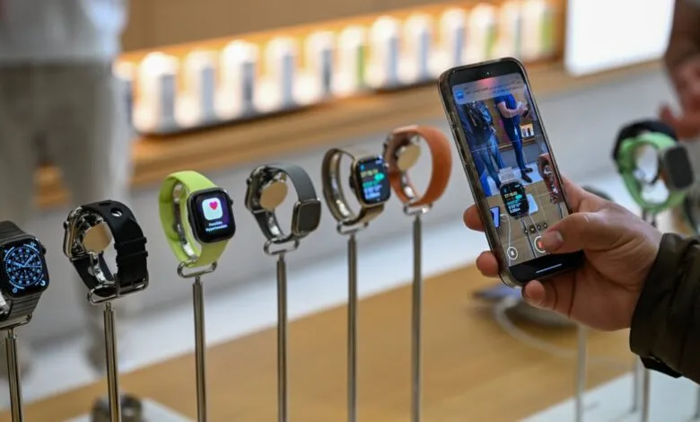 Hand holding a smartphone recording a display of various Apple Watches with different colored bands.