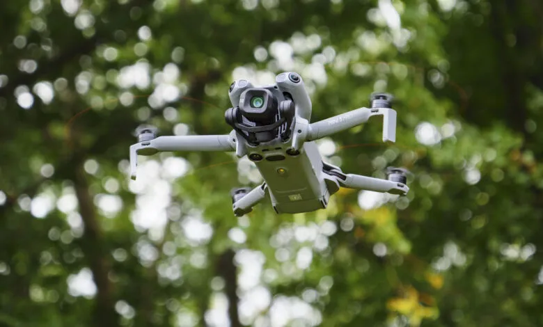 A DJI Mini 5 Pro drone in flight against a blurred green background of trees.