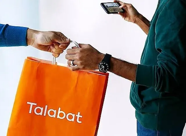 Delivery person handing an orange Talabat bag to a customer, who is using a smartphone.