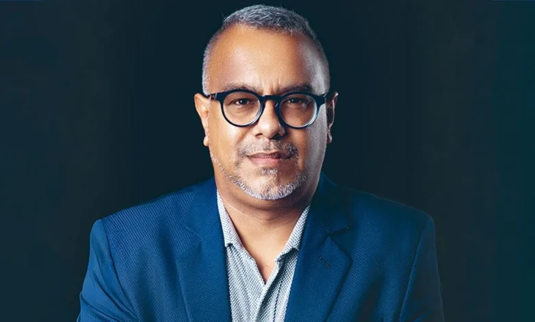 Headshot of a man in a blue blazer, wearing glasses, against a dark background.