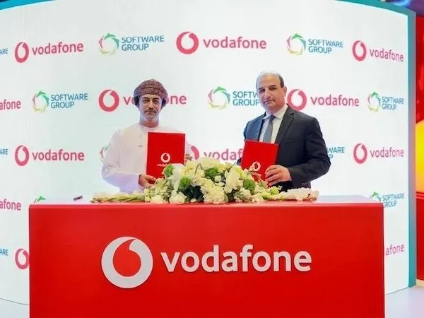 Two men in business attire sign a Vodafone agreement, standing behind a red table with the Vodafone logo.