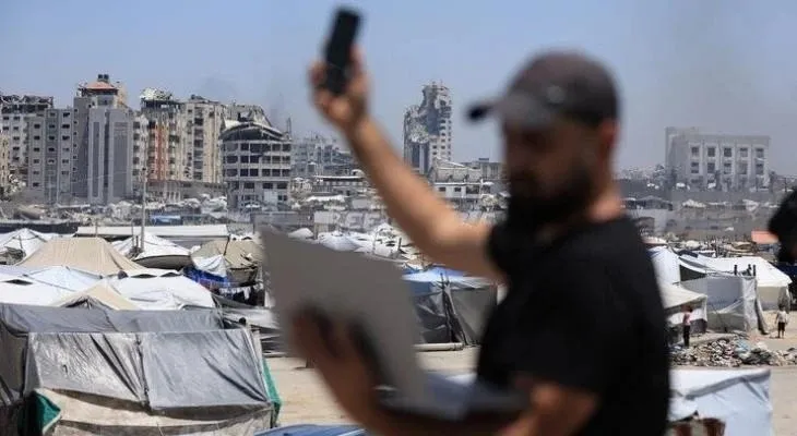 Person using phone and laptop in front of war-damaged buildings and refugee tents.
