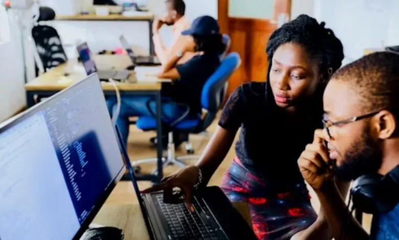 Two colleagues collaborate on a laptop, reviewing code on a large monitor in a busy office.